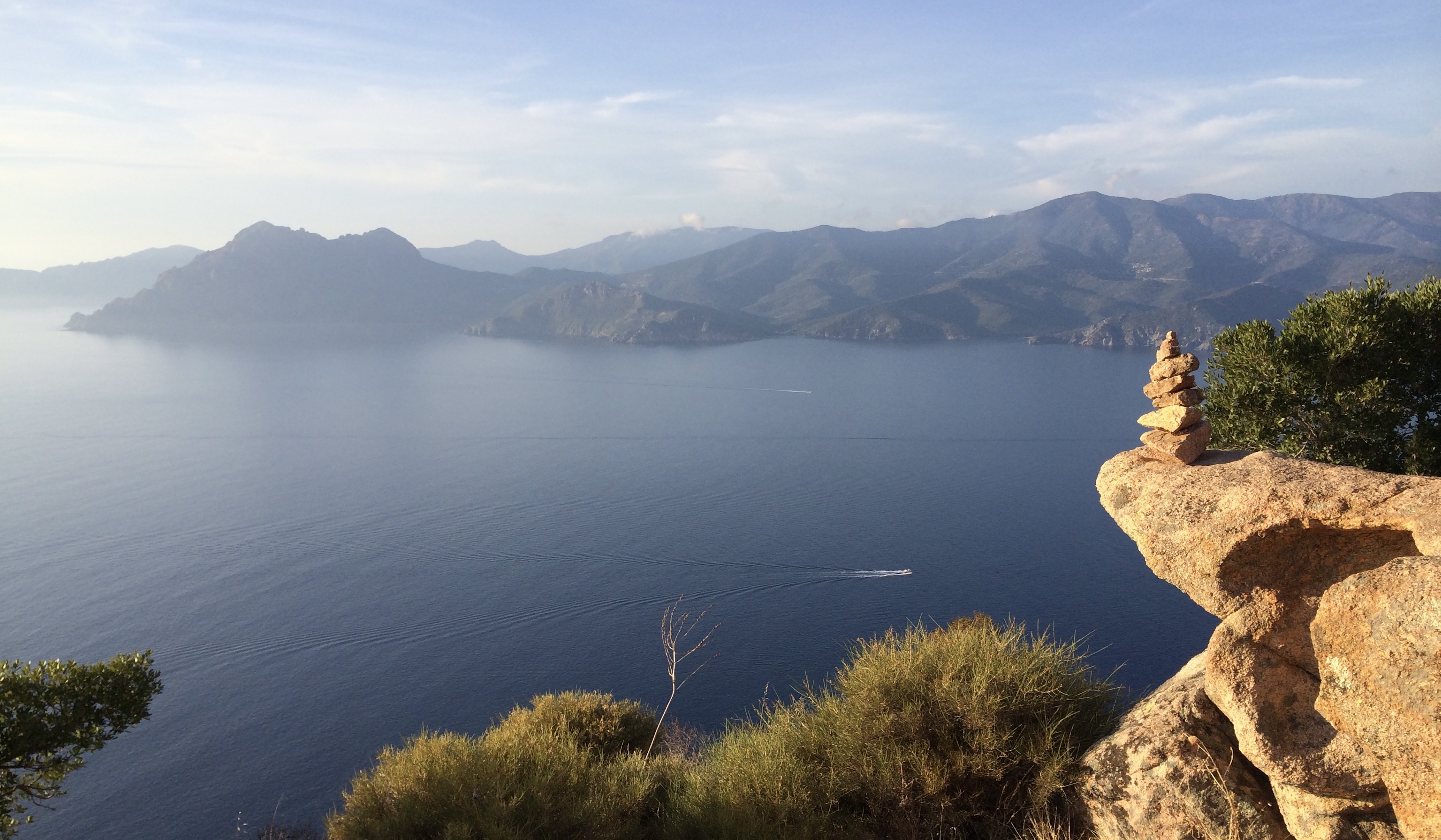 Scenic view of the sea from a rocky cliff, with a mountain range in the background and a small boat on the water.